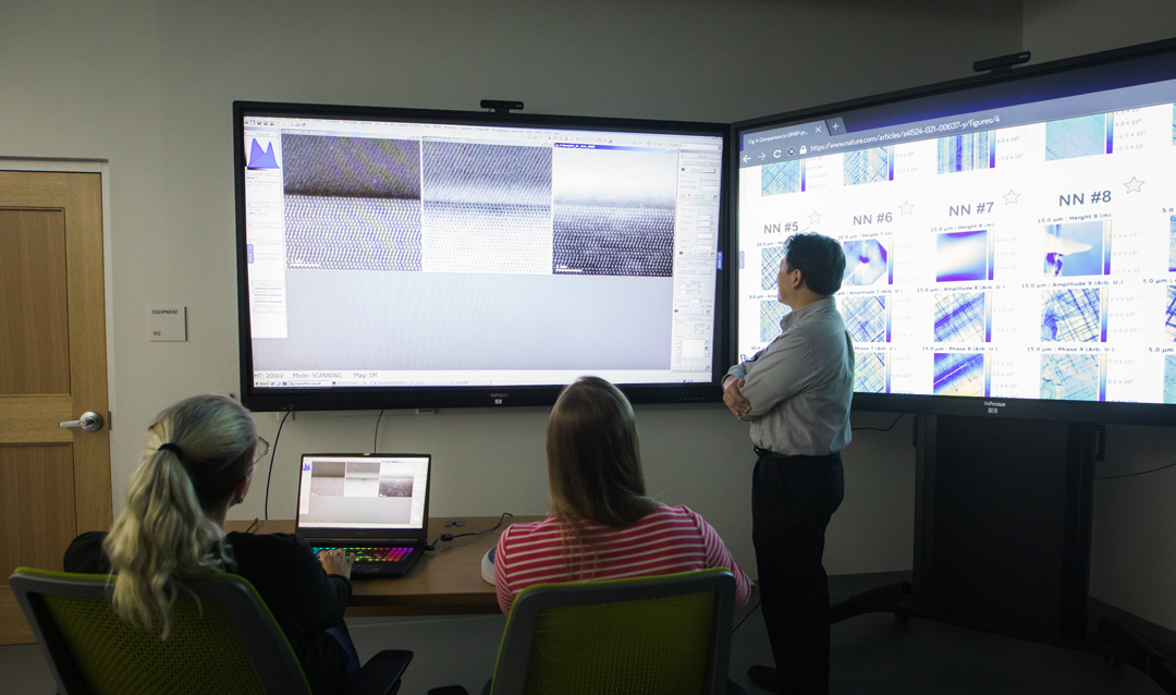 Two women sit at a table controlling a microscope remotely while a man stands next to a large touchscreen.