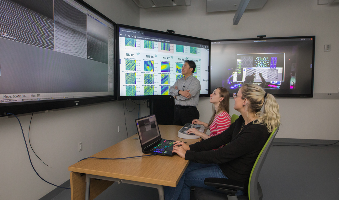 Two women sit at a table controlling a microscope remotely while a man stands next to a large touchscreen.