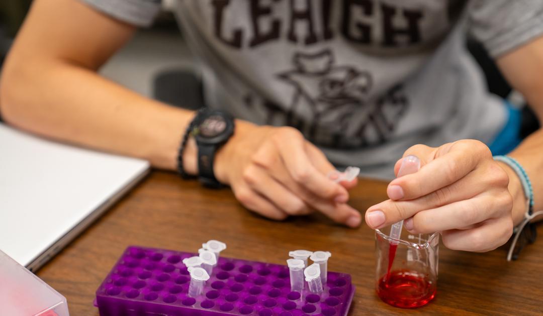 Person handling test tubes with red liquid at a lab table.