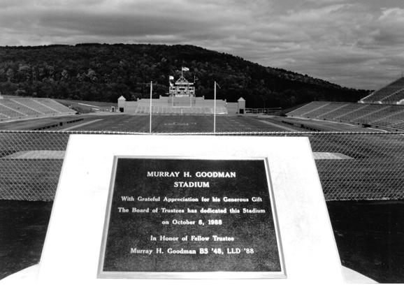 A black-and-white photo of a dedication plaque with a stadium in the background.