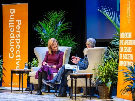 Arianna Huffington engaged in discussion with President Joseph J. Helble on stage, surrounded by plants and banners.