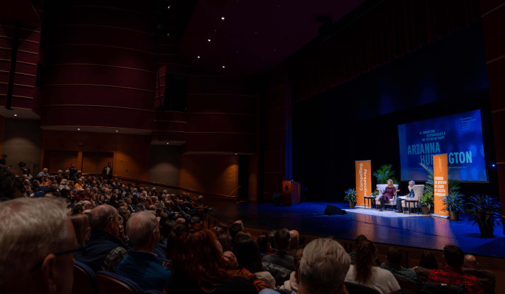 A crowded theater with an audience facing a stage featuring Arianna Huffington and President Joseph Helble and bright lighting.