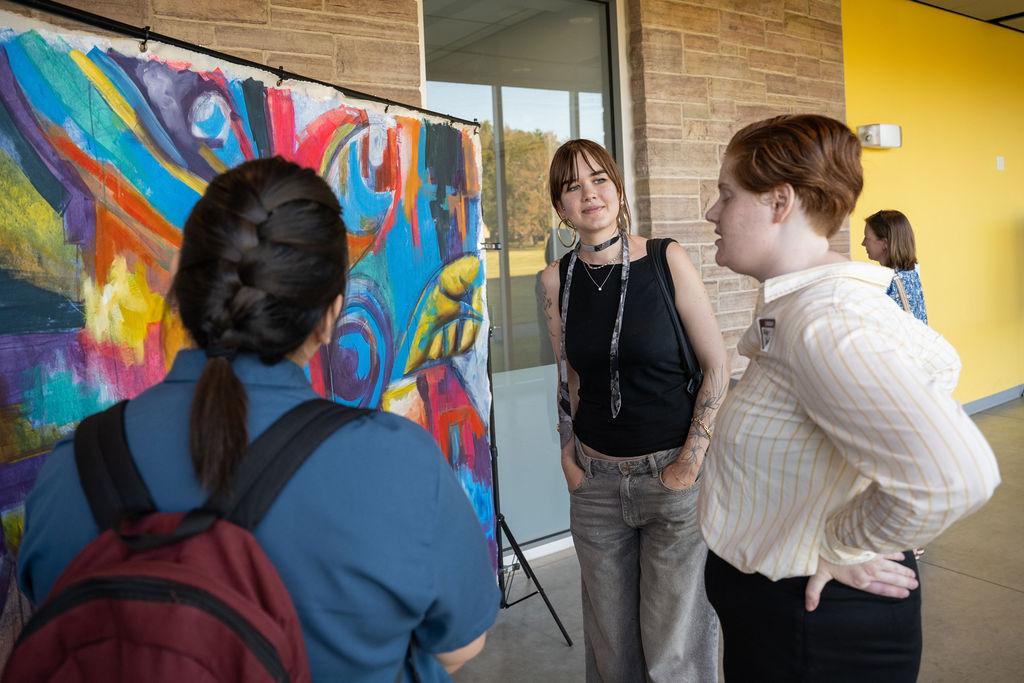Three people engage in conversation by a colorful abstract painting in a gallery.