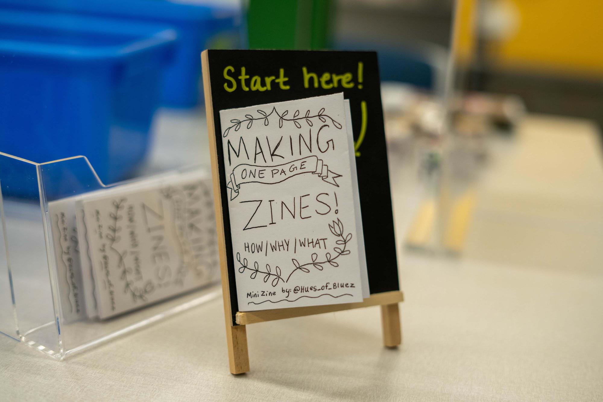 A small chalkboard sign displaying instructions for making zines, with a stack of zine booklets nearby.