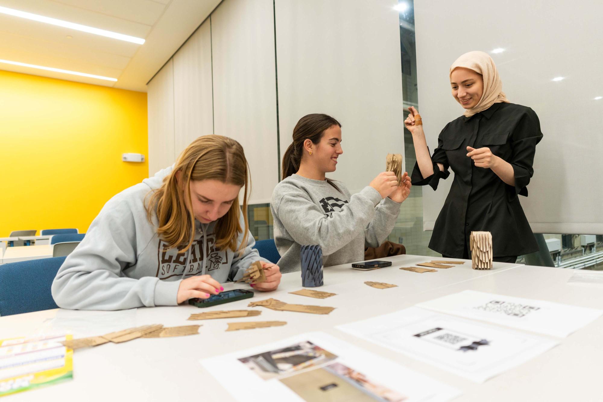 Three women engage in an activity at a table, with one focused on a phone and others holding items.