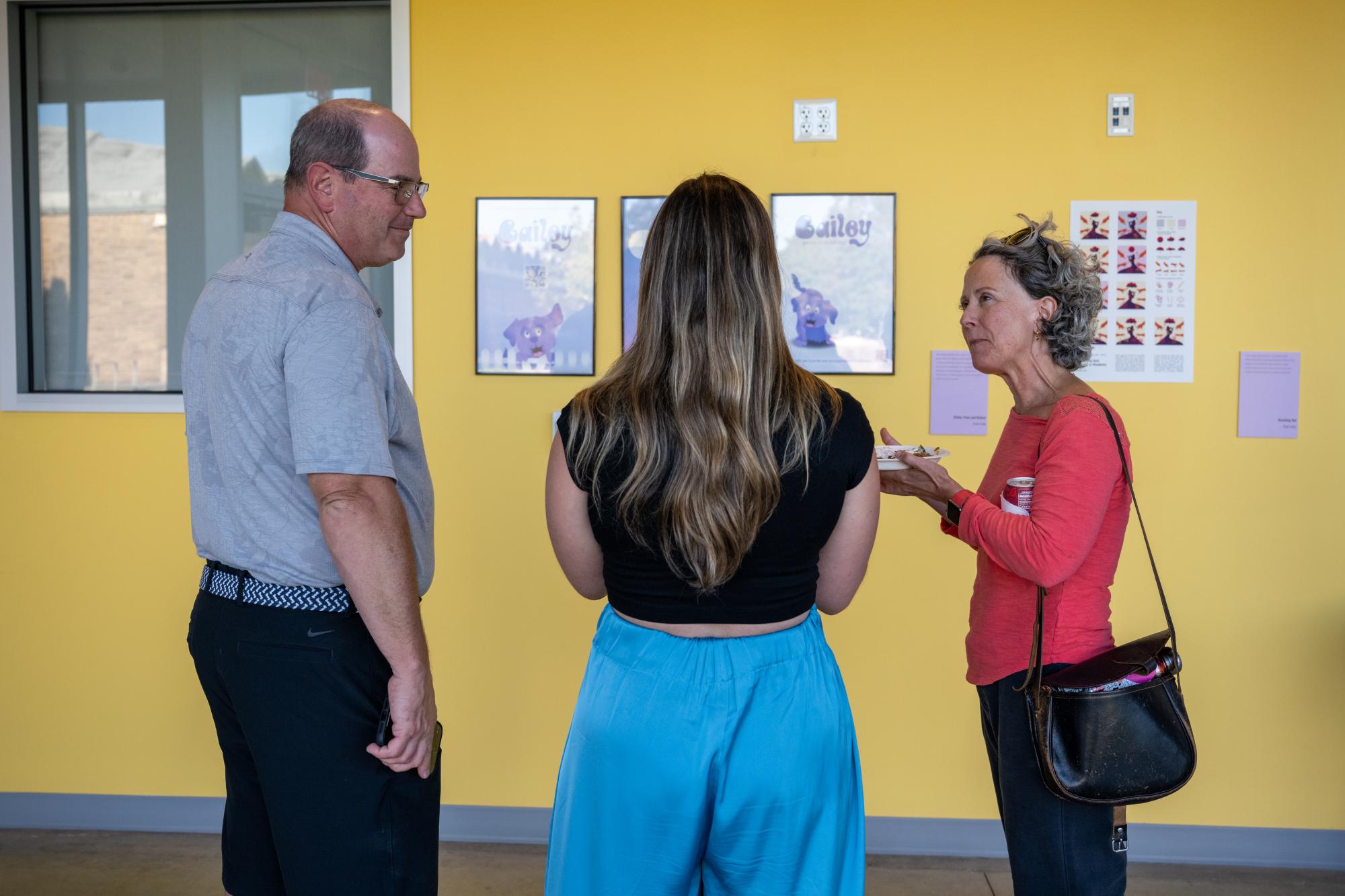 Three people converse in front of artwork on a yellow wall in a bright room.