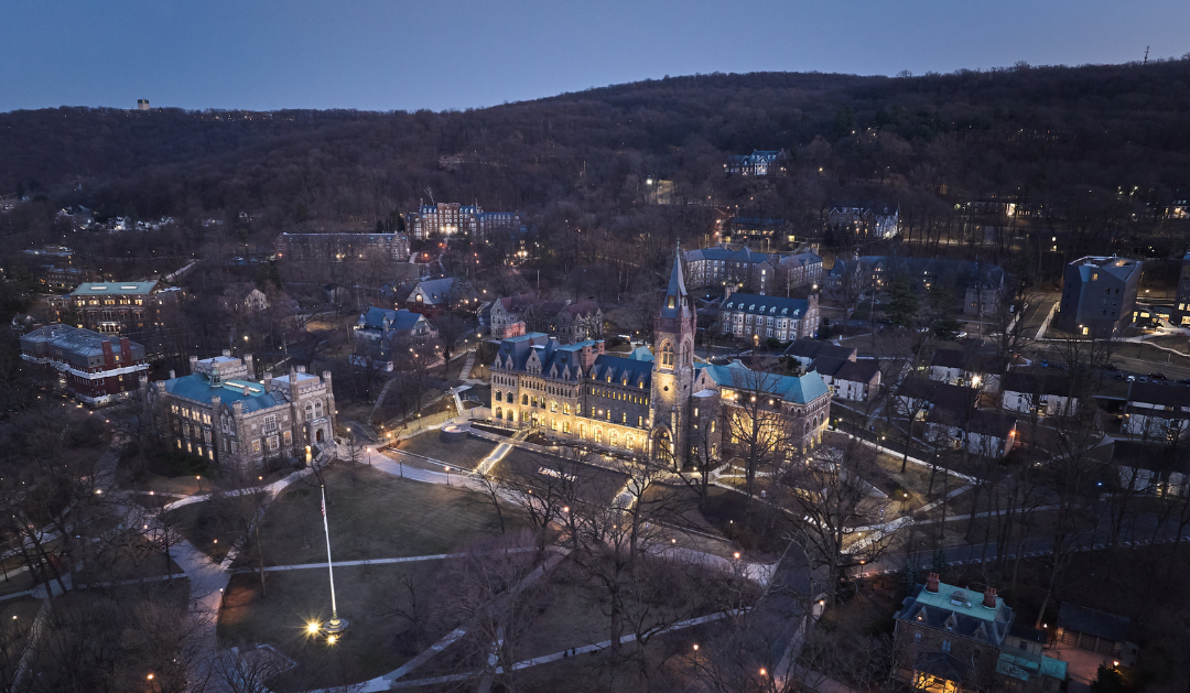 Aerial view of a campus at twilight, buildings illuminated, surrounded by trees and hills.