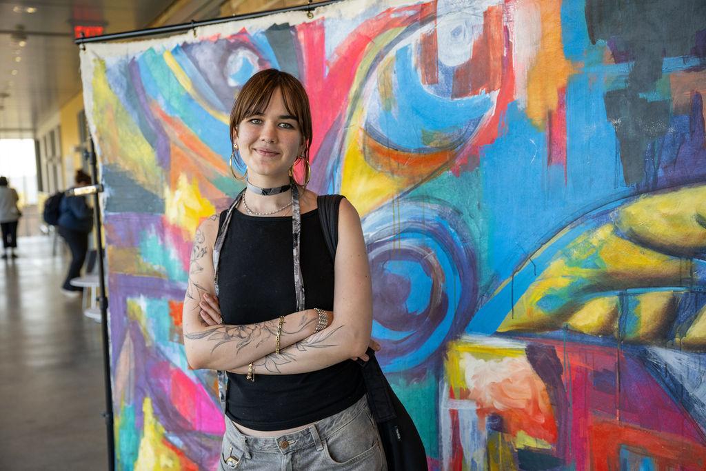 A woman with short hair and tattoos stands confidently in front of a colorful painted mural she created.