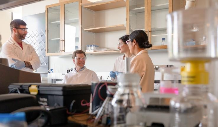 Four scientists in lab coats discuss in a laboratory, with equipment in the foreground.