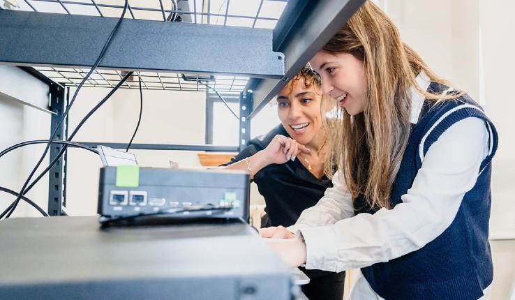 Two women focused on a device in a tech setting, smiling and collaborating.