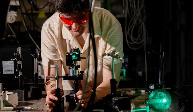 A researcher in red safety glasses adjusts equipment in a dark laboratory with green lighting.