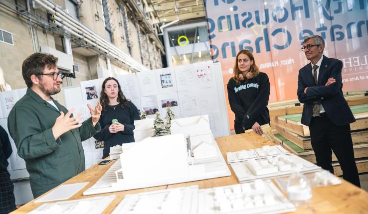 A group discusses architectural models in a workshop, with one person presenting.