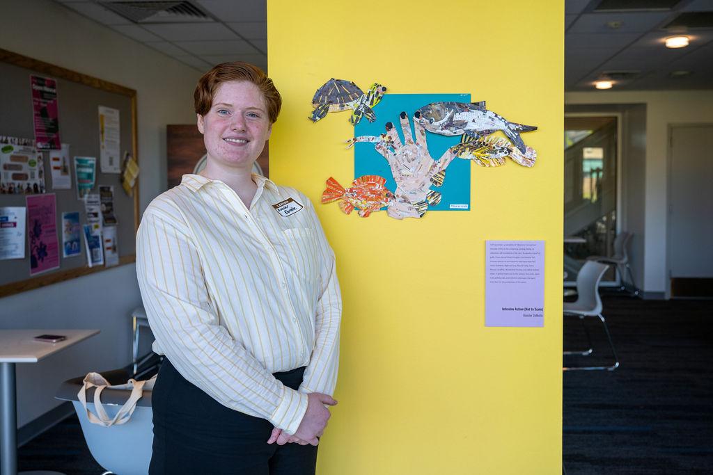 Woman in a striped shirt stands beside mixed media collage on a yellow wall.