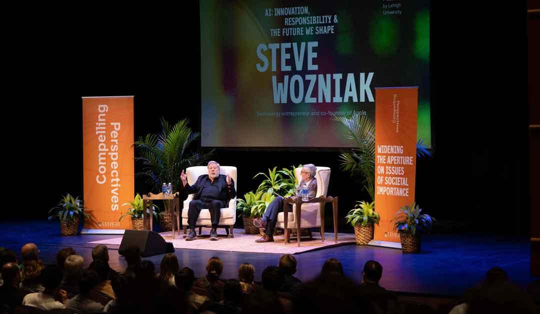 Steve Wozniak speaking on stage, seated with President Joseph J. Helble, surrounded by an audience.