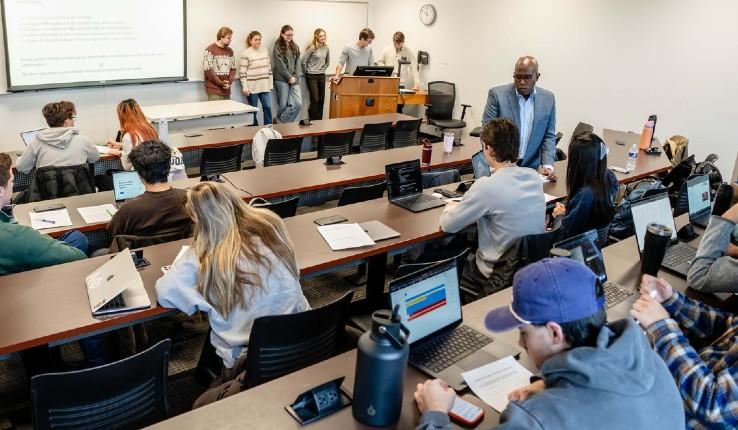 Students seated at desks in a classroom, with some presenting at the front.