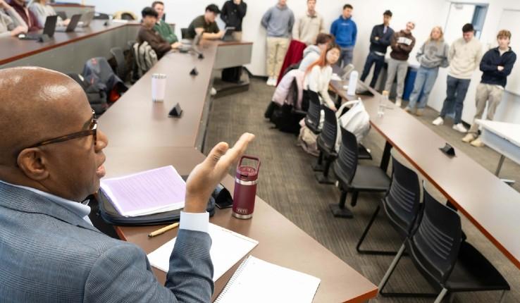 A speaker gestures while addressing an audience in a classroom setting.