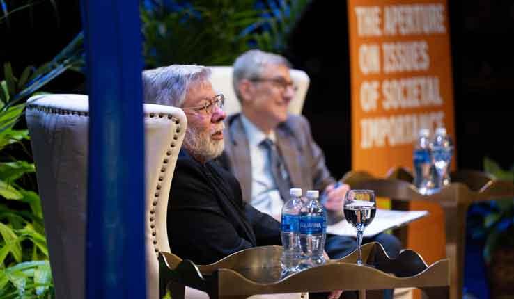 Two men seated on stage during a discussion, surrounded by plants and water bottles.