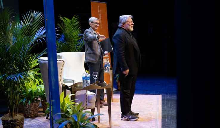 Two speakers stand on stage, surrounded by greenery and facing the audience.