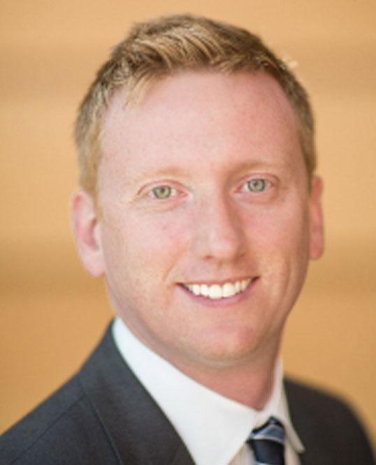 Smiling man in a suit with short blonde hair, warm background.