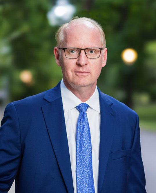 Man in a blue suit and glasses standing outdoors, with blurred greenery and lights in background.