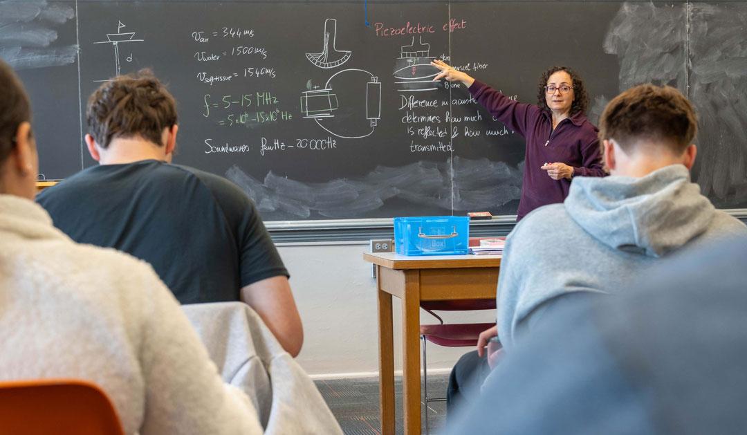 A teacher gestures toward a chalkboard while students sit at desks, focused on the lesson.