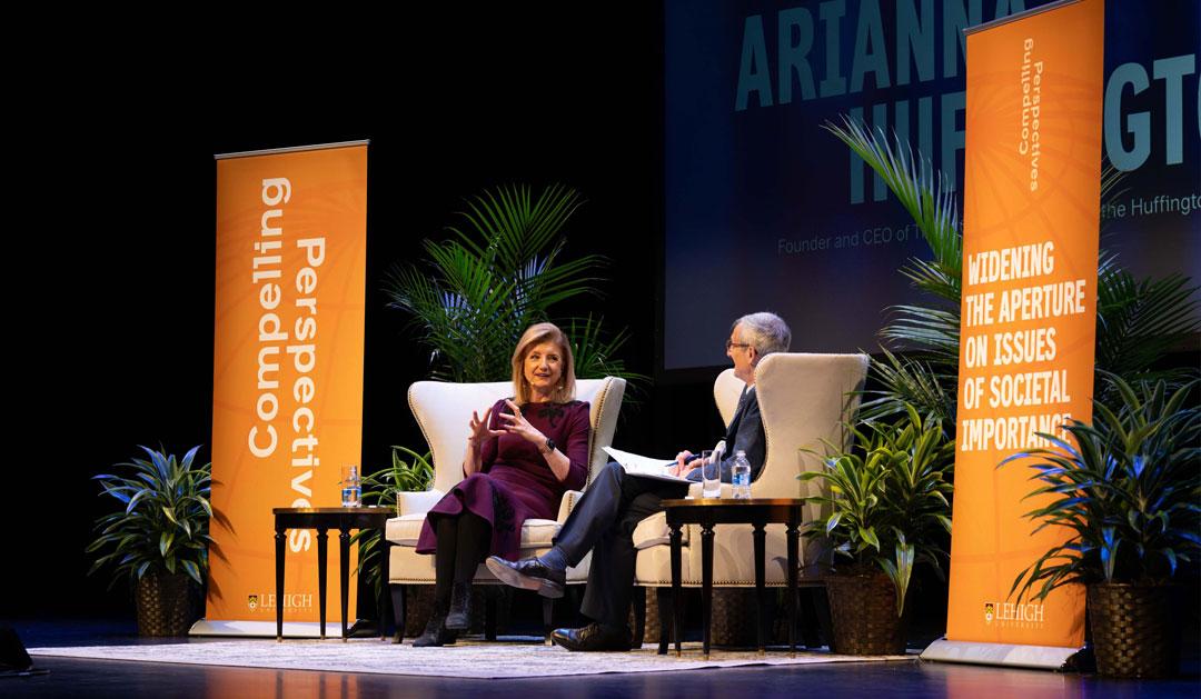 Two speakers engage in conversation on stage, flanked by decorative plants and banners.