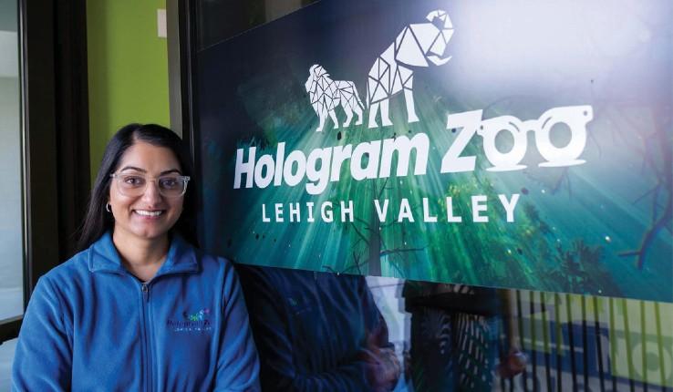 A woman smiles beside a sign for Hologram Zoo, Lehigh Valley, featuring animal silhouettes.