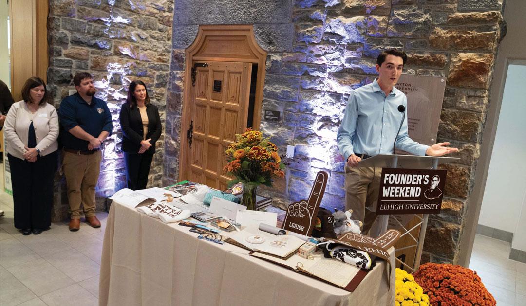 A speaker presents at an event with a table of displays and attendees in the background.