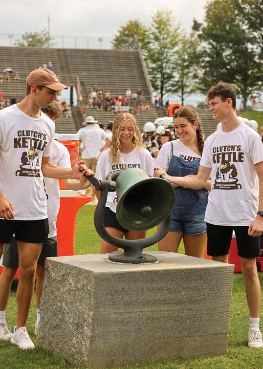 Four people gather around a large bell on a stone pedestal at an outdoor event.