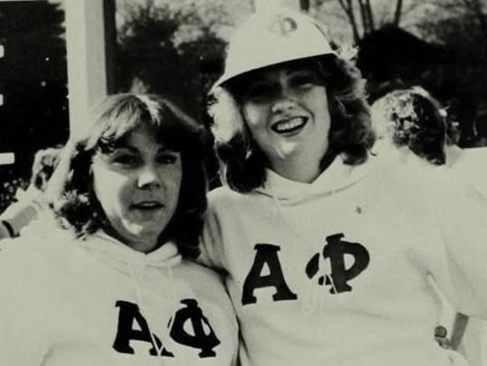 Two women in matching white sweatshirts with Greek letters, smiling together outdoors.