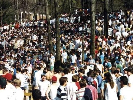 A large crowd of people gathers in a wooded area, with many wearing casual attire.
