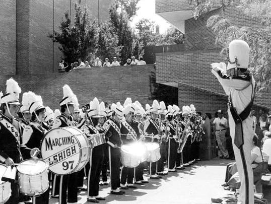 A marching band in uniform performs, led by a conductor in front of a crowd. Black and white.