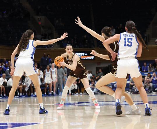 A basketball player in brown prepares to pass while two defenders in white guard her.