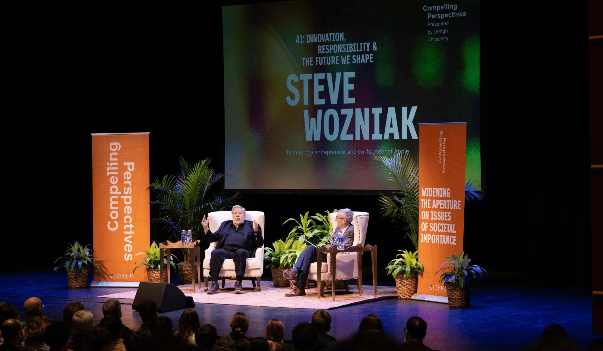 Steve Wozniak speaking on stage, seated with President Joseph J. Helble, surrounded by an audience.