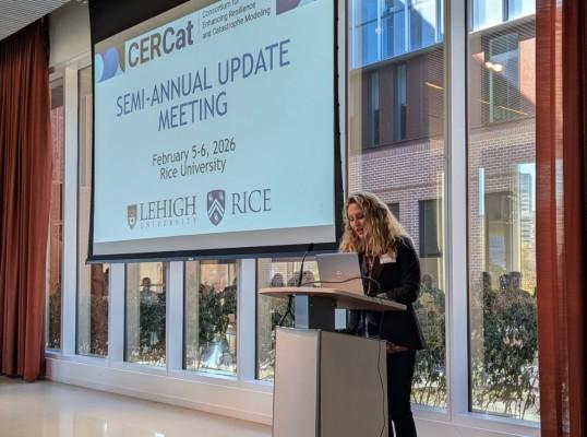 A speaker at a podium presents during a meeting, with large windows in the background.
