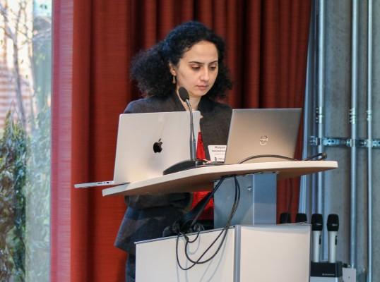 A woman with curly hair stands at a podium, focused on a laptop in front of her.