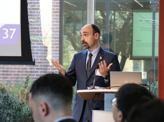 A man in a suit gestures while speaking at a podium in a bright, modern setting.