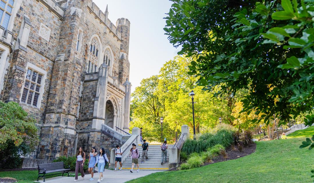 Students walk past a grand stone university building on a sunny day.
