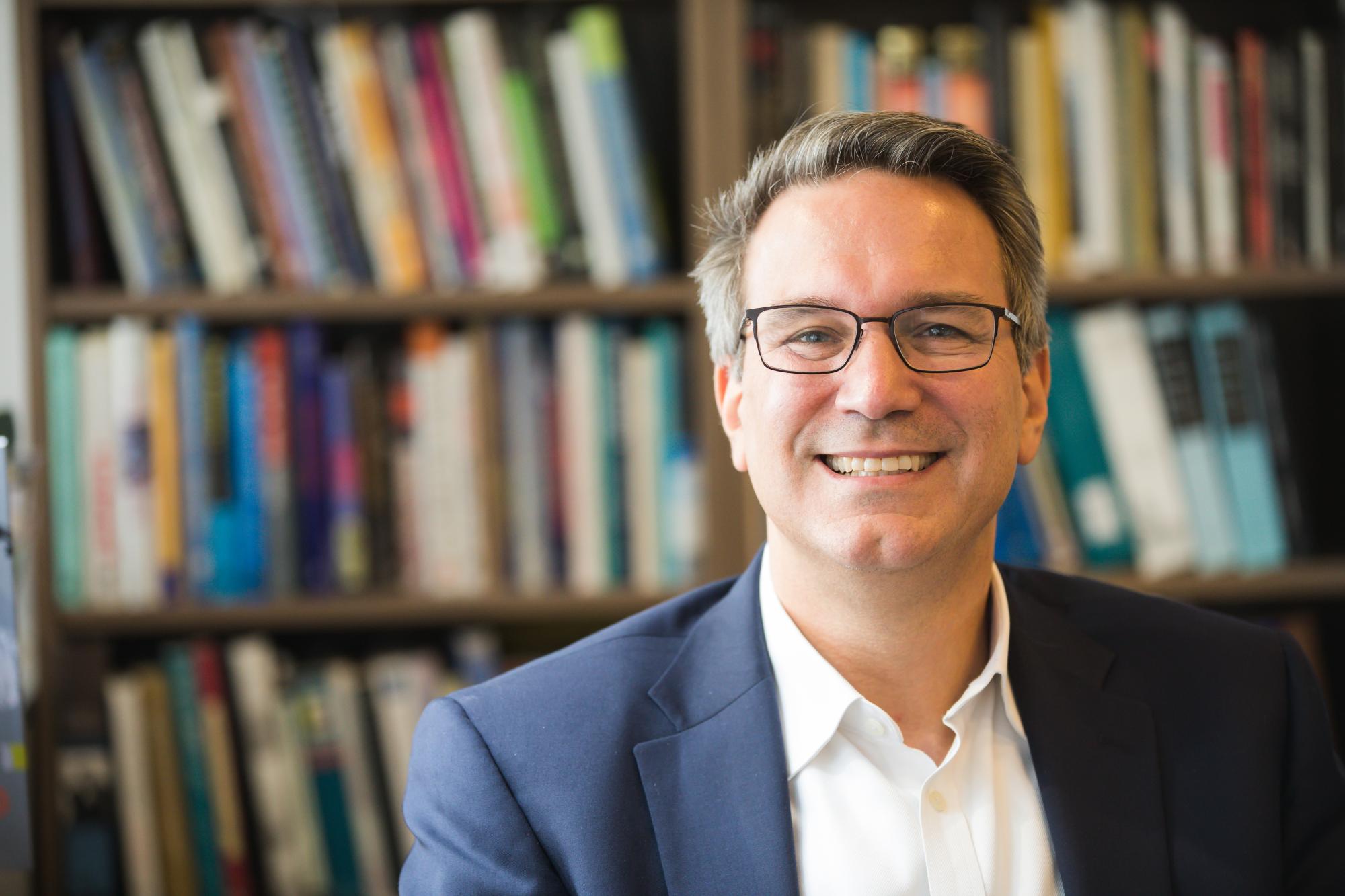 Man with glasses smiling, bookshelf in the blurred background.