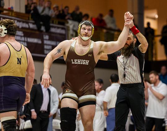 Wrestler in brown 'LEHIGH' singlet celebrates win, arm raised by referee.