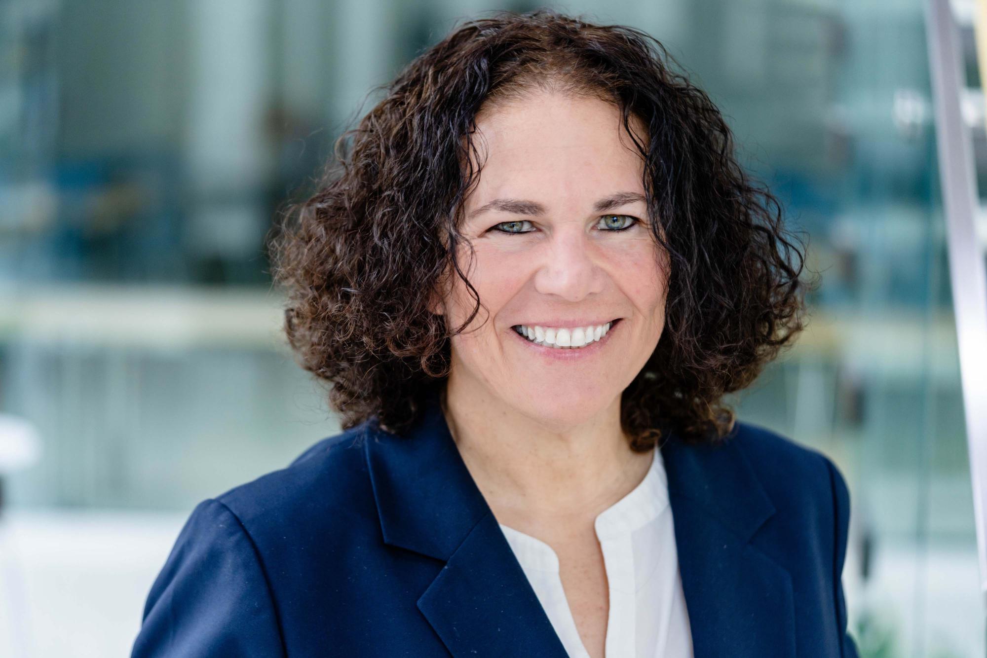 Smiling woman with dark curly hair in a navy blazer and white shirt.
