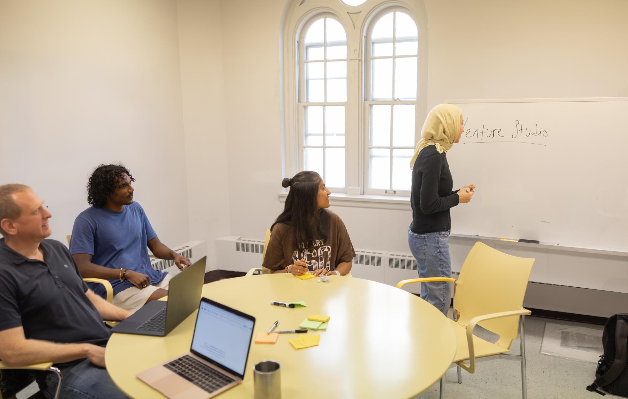 Four people meet in a bright room. One writes on a whiteboard, three sit at a round table.