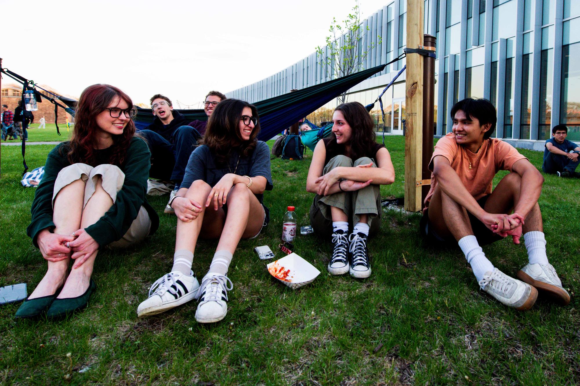 Photograph of students laughing while sitting on a grassy lawn near a campus building.