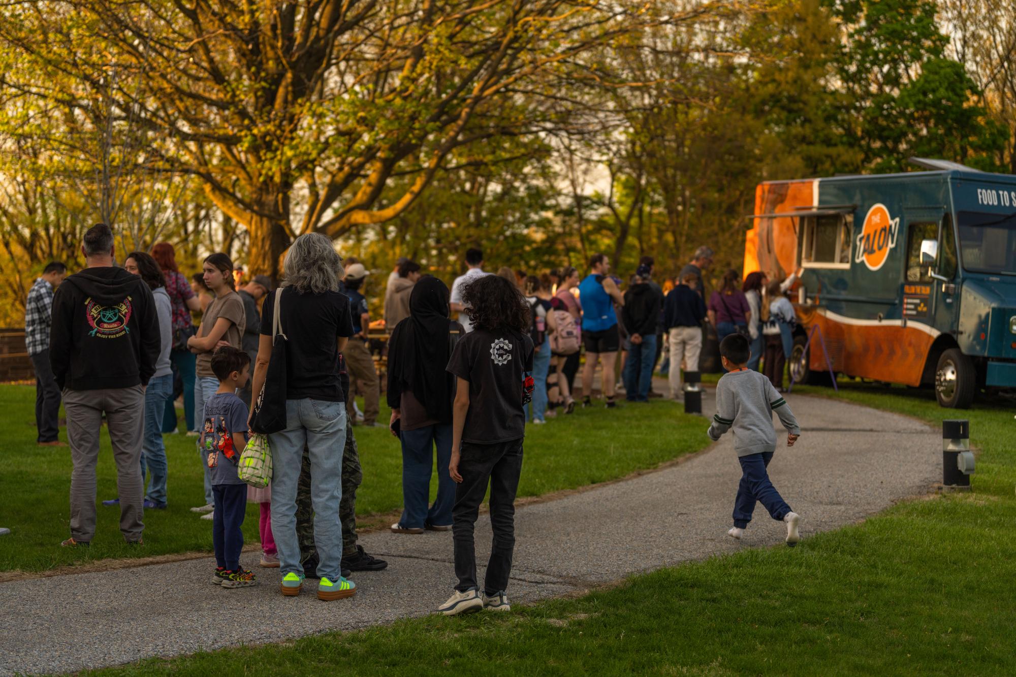 Photograph of a crowd waiting for a teal and orange food truck in a park at sunset.