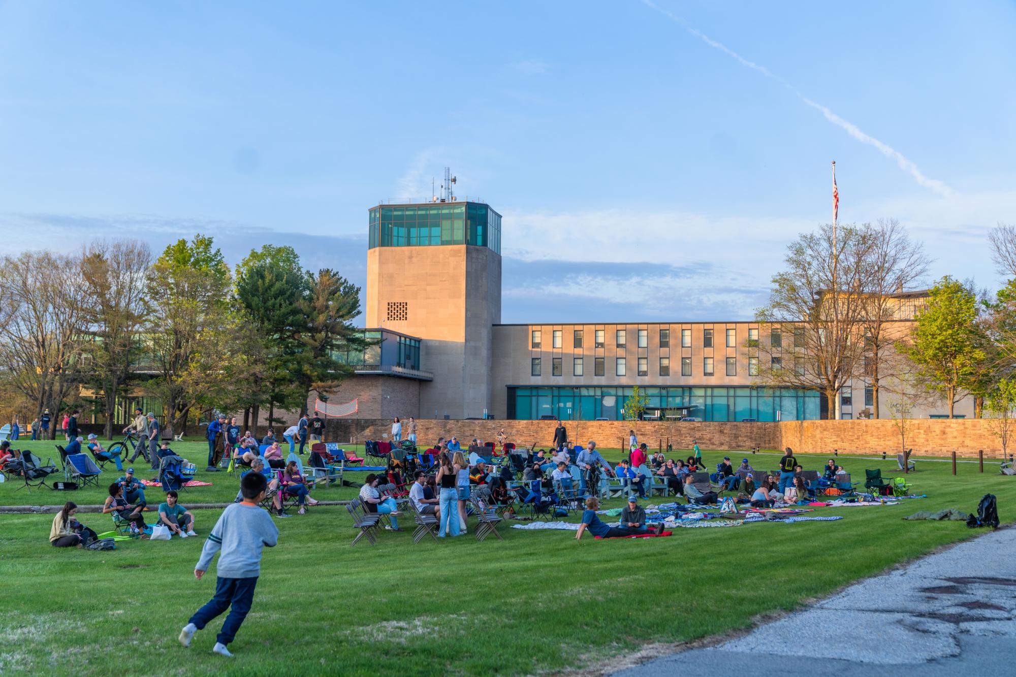 Photograph of people lounging on a grassy lawn in front of a modern stone building and tower.