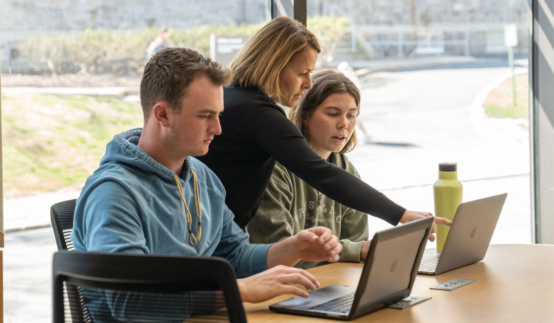 Woman points to laptop screen while two students work on their computers.