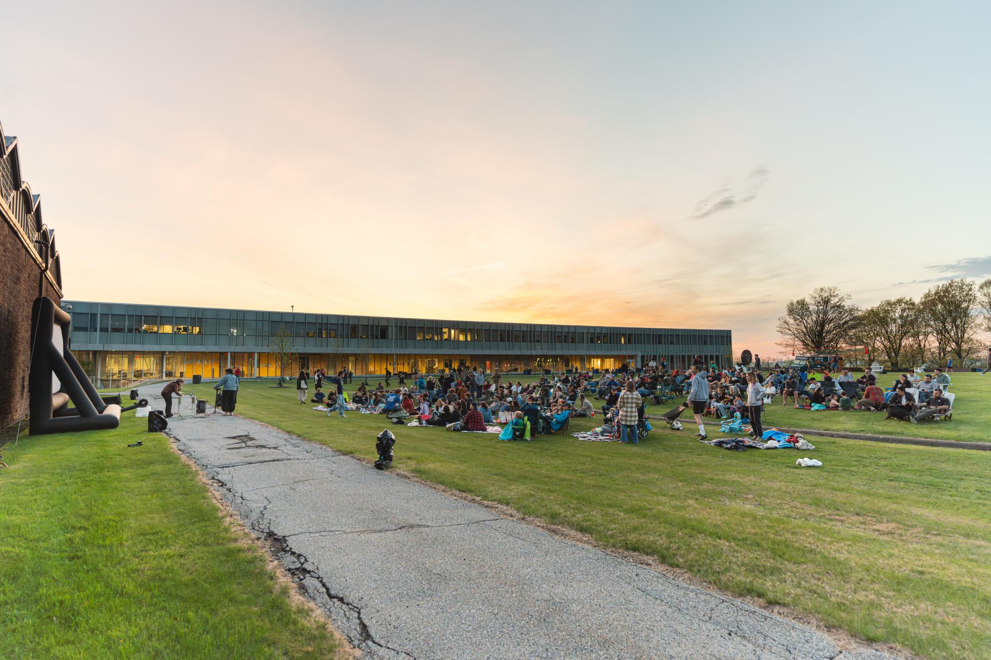 Photograph of a crowd gathered on a lawn at sunset in front of a long modern building.