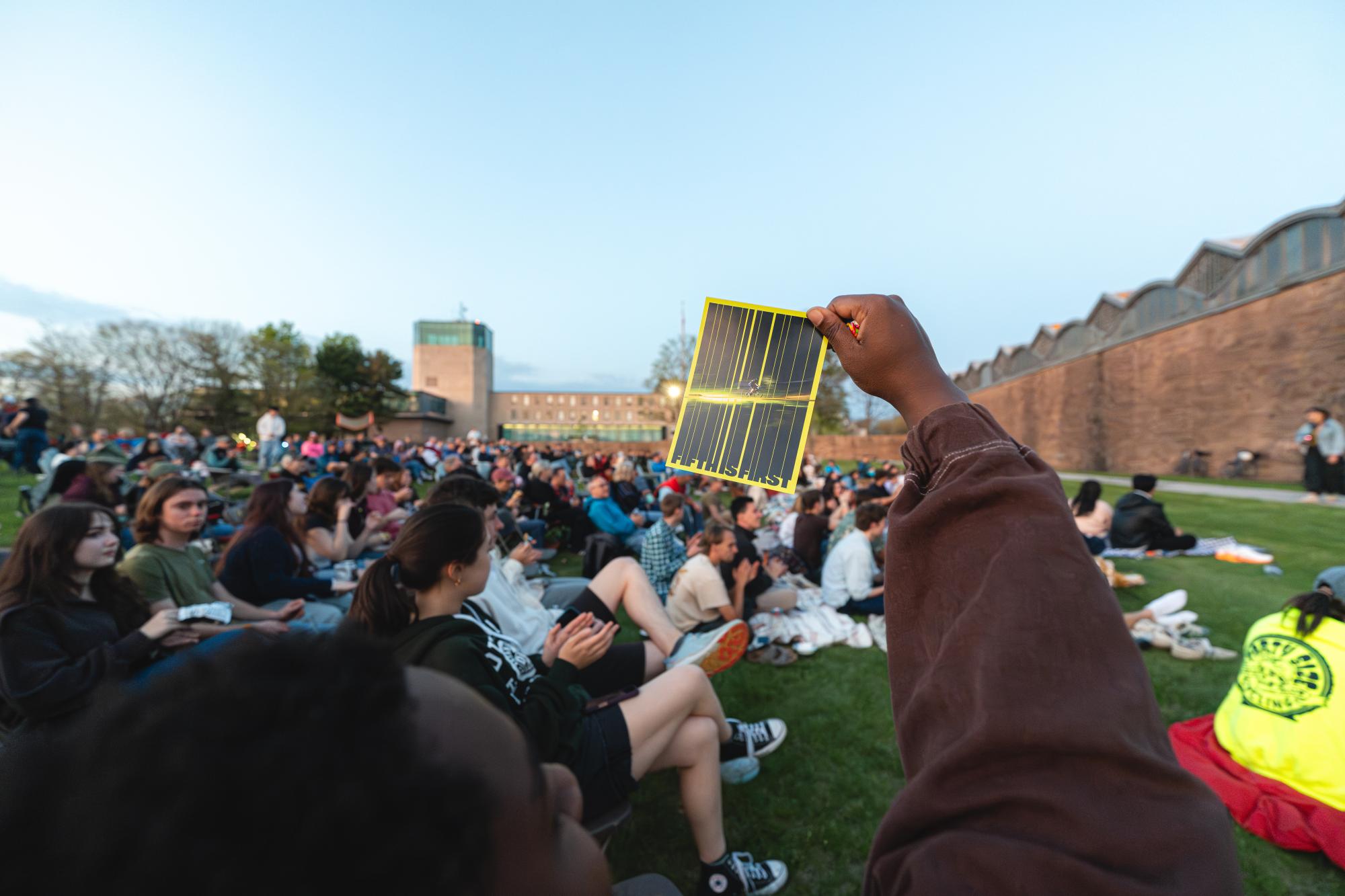 Photograph of a hand holding a solar viewer before a crowd sitting on a lawn.