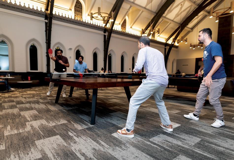 Four men playing ping pong in a spacious room with high ceilings and arched windows.