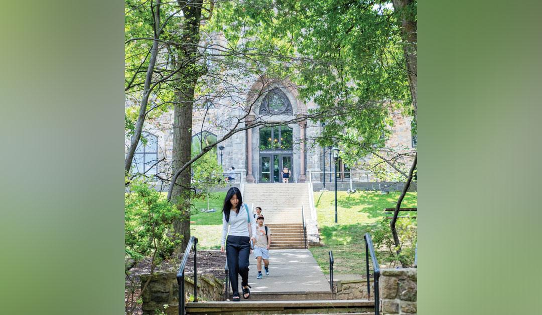 Students walking down stone steps from an arched stone building, sunny day.